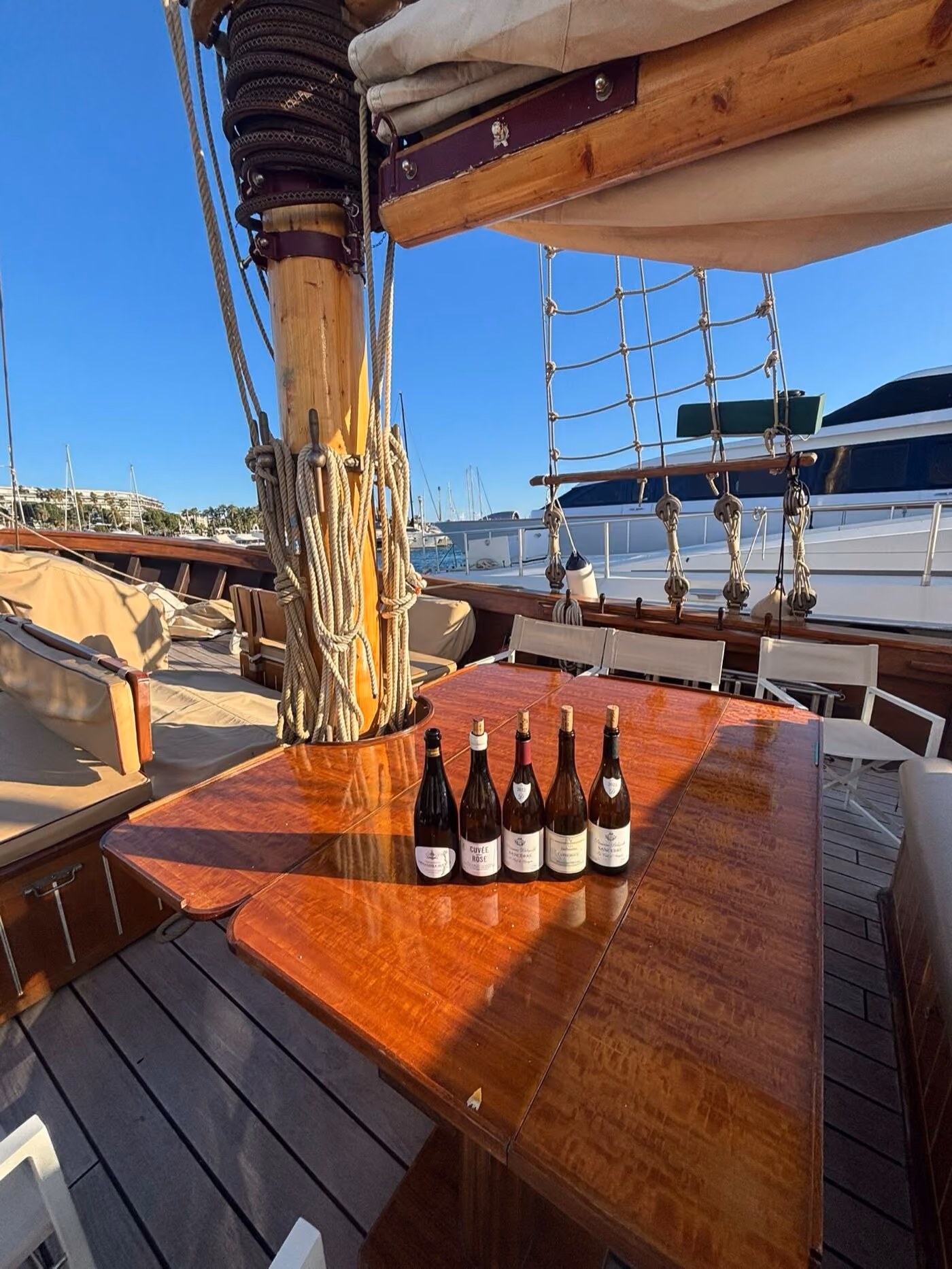 Cinq bouteilles alignées sur la table en bois verni d'un voilier, gréement et port en toile de fond