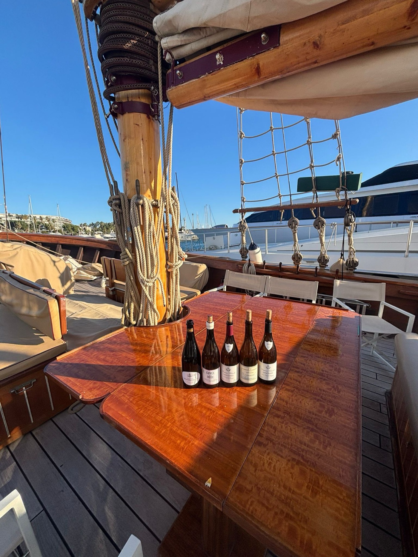 Five bottles lined up on the varnished wooden table of a sailboat, rigging and marina in the background