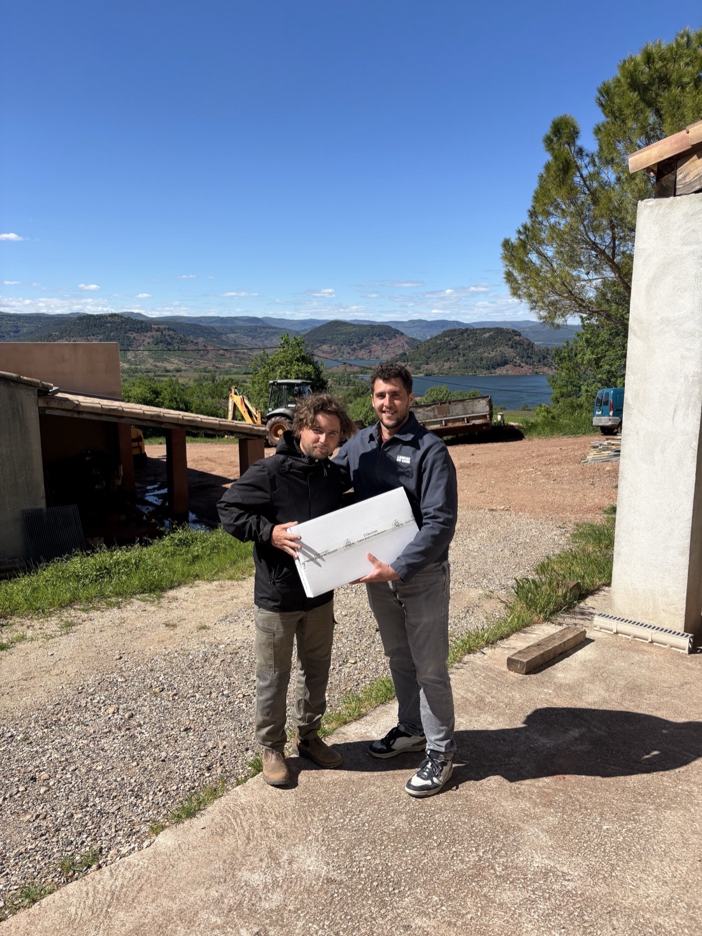 Guillaume y Florent posando con una caja de L'Oratoire Saint Jean d'Aureilhan ante las colinas y el lago Salagou